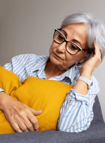 An older woman wearing glasses is sitting by herself, holding a pillow. She looks sad.